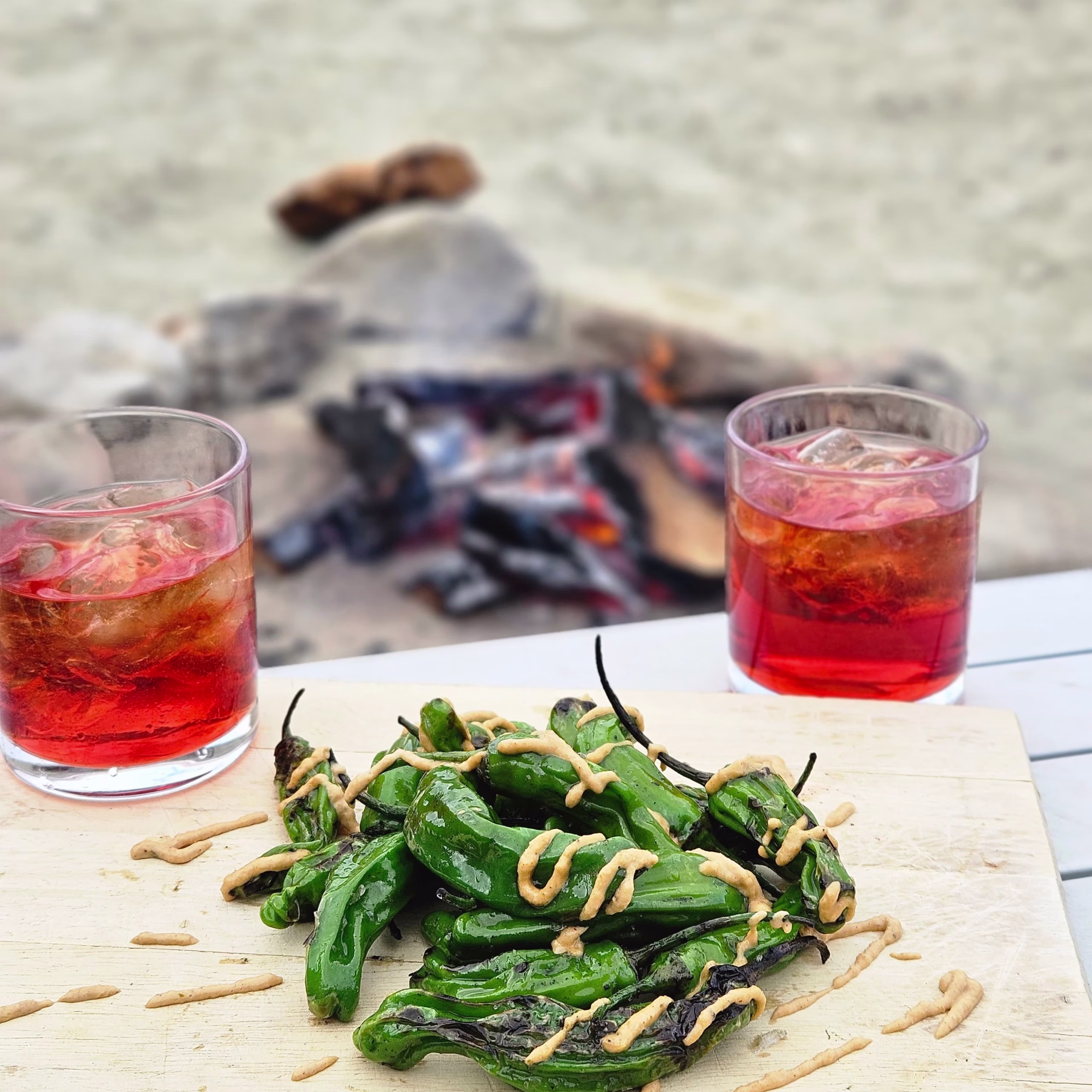 Roasted shishito peppers served on a board with cocktails at a campsite.